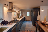 A cool, spacious Pantry blue Shaker kitchen features a long sink and cooker run with a Carrara marble worktop, a bank of appliance cabinets, and a pantry larder. Lovingly decorated with mini creamware wall lights, a laundry maid, a large pleated porcelain pendant light, houseplants, a charcuterie board, and fresh vegetables, it creates a humble, practical kitchen space.