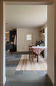 Looking through an open corridor lies a cool, spacious Pantry blue Shaker kitchen featuring a bank of appliance cabinets and a pantry larder. Beside it is a small dining area with antique wooden tables and chairs. Lovingly decorated with a large rug, red detailed cloth, houseplants, and fresh vegetables, it creates a humble, practical kitchen and dining space.