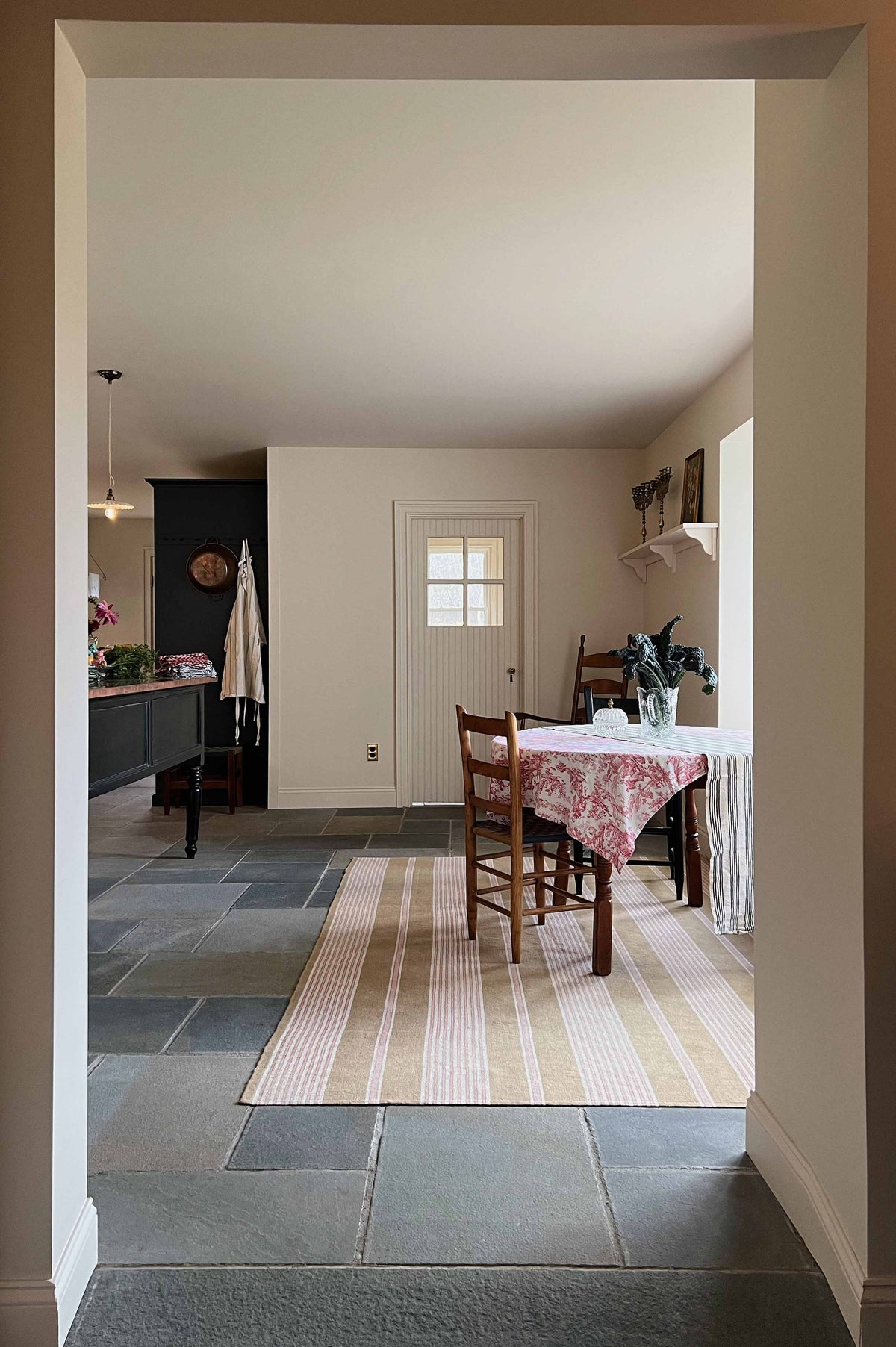 Looking through an open corridor lies a cool, spacious Pantry blue Shaker kitchen featuring a bank of appliance cabinets and a pantry larder. Beside it is a small dining area with antique wooden tables and chairs. Lovingly decorated with a large rug, red detailed cloth, houseplants, and fresh vegetables, it creates a humble, practical kitchen and dining space.