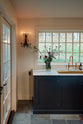 A quiet corner of a cool, spacious Pantry blue Shaker kitchen features a long sink and cooker run with a Carrara marble worktop, and an aged brass ionian tap. Lovingly decorated with a large vase of wild flowers and an antique wall candle holder, it creates a unique and practical kitchen space.