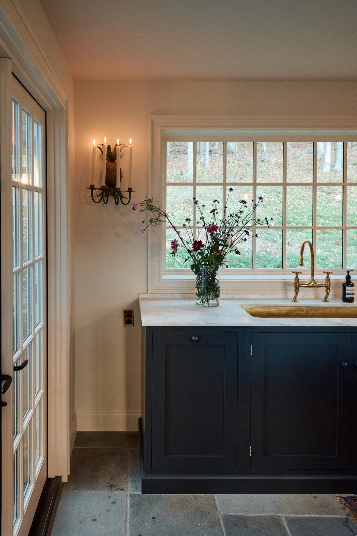 A quiet corner of a cool, spacious Pantry blue Shaker kitchen features a long sink and cooker run with a Carrara marble worktop, and an aged brass ionian tap. Lovingly decorated with a large vase of wild flowers and an antique wall candle holder, it creates a unique and practical kitchen space.
