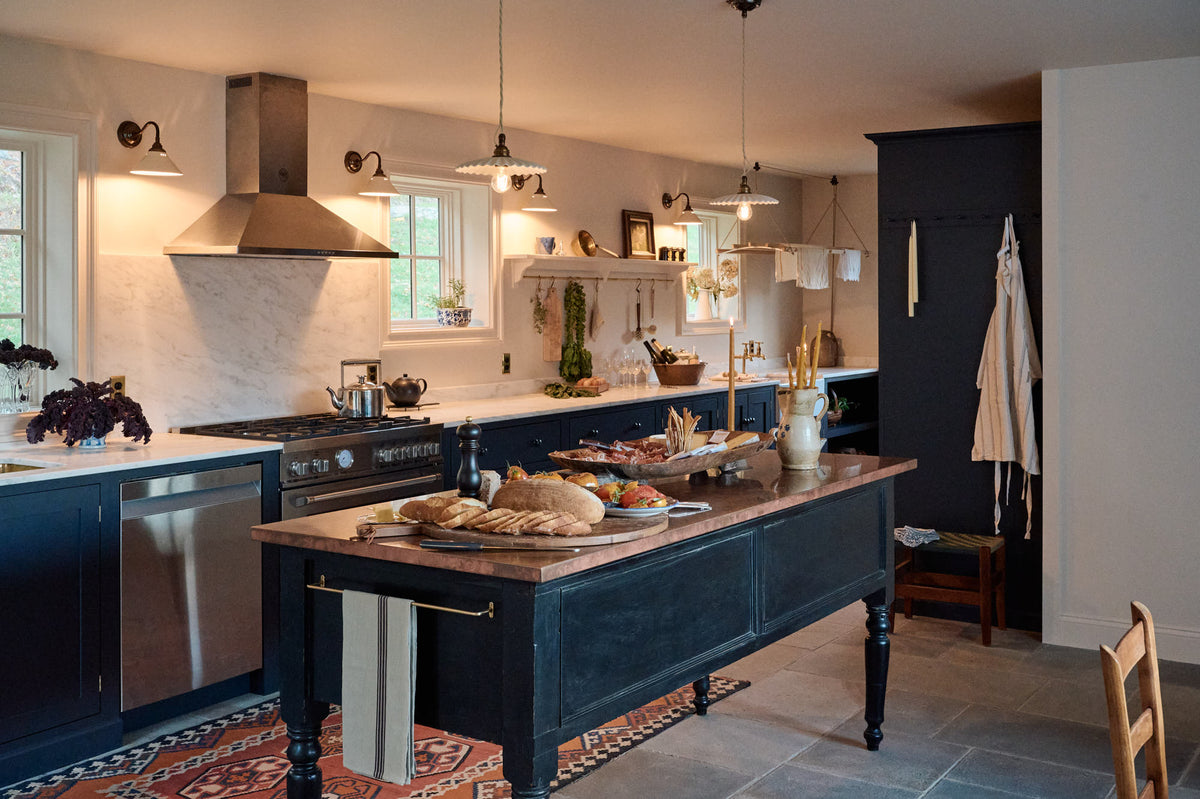 A cool, spacious Pantry blue Shaker kitchen features a long sink and cooker run with a Carrara marble worktop. Lovingly decorated with mini creamware wall lights, a laundry maid, a large pleated porcelain pendant light, houseplants, and candles, it creates a humble, practical kitchen space.