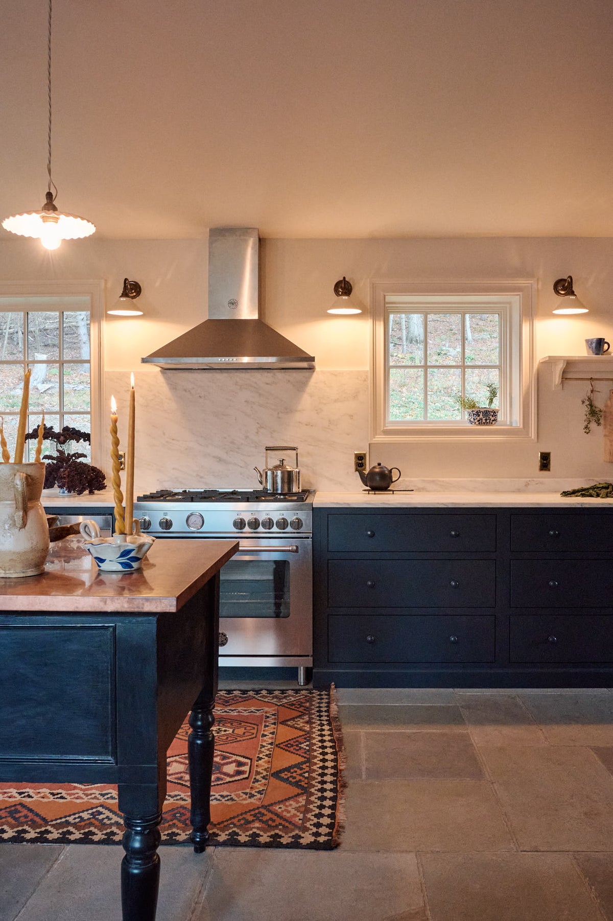 A cool, spacious Pantry blue Shaker kitchen features a long sink and cooker run with a Carrara marble worktop. Lovingly decorated with mini creamware wall lights, a laundry maid, a large pleated porcelain pendant light, houseplants, and candles, it creates a humble, practical kitchen space.