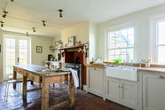A small and quaint mushroom-coloured Shaker kitchen features a sink run made up of a mix of stave hardwood and Carrara marble worktops, a shelf, and a black stove nestled inside the original building’s chimney breast. Beautifully decorated with vintage furniture such as a large solid oak table, a silver scale, and assorted bric-a-brac, it creates a lovely social space full of character.