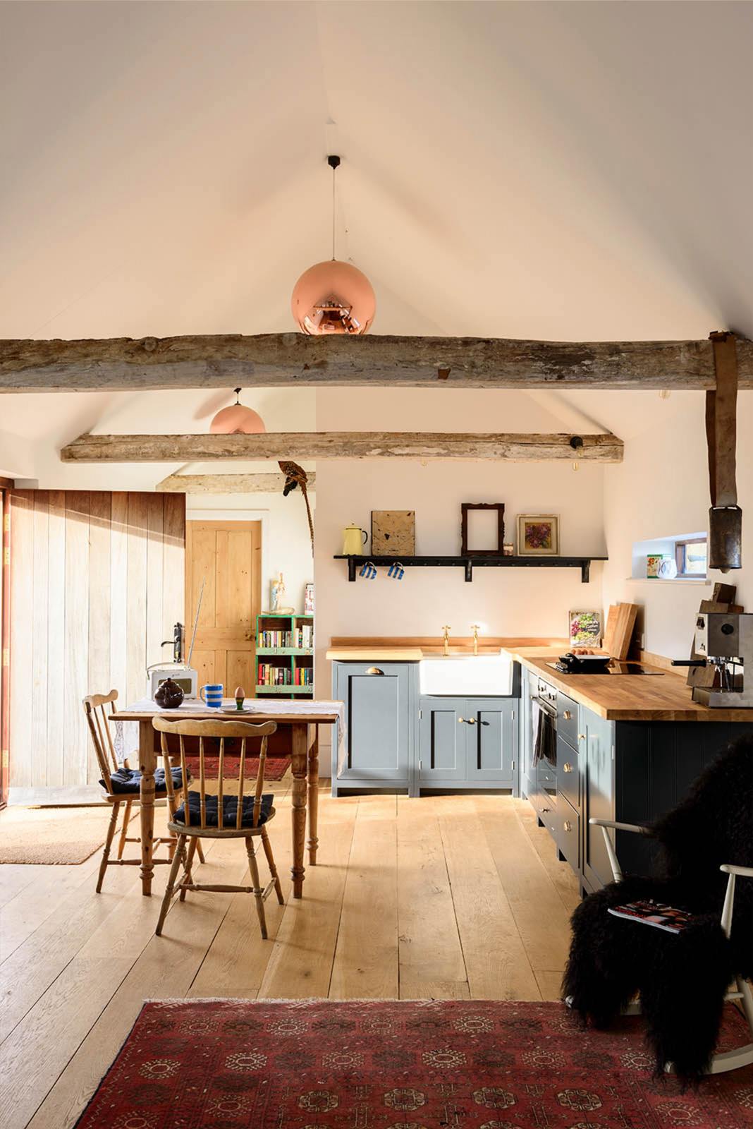 A small and humble pantry-blue Shaker kitchen features an L-shaped sink and cooker run with a stave hardwood worktop, a single Belfast sink, and aged brass Mayan taps. Beautifully decorated with a taxidermy bird sitting on the roof’s wooden beams, vintage furniture, books, and assorted bric-a-brac, creates a heartwarming family farm kitchen space.