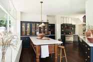 A large pantry blue shaker kitchen features a sink and cooker run with Carrara marble worktop, countertop cupboards with Scullery yellow interior, and a wide Heirloom dairy table. Beautifully decorated with artisan ceramics, copper pans, deVOL Heirloom gaselier lights, a pot hanger, an aged brass pot filler tap, and Helen Stools, the kitchen presents a thoughtfully curated mix of collected pieces that fit together effortlessly.