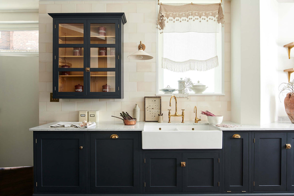 A large pantry blue shaker kitchen features a sink and cooker run with Carrara marble worktop, a double Belfast sink, an aged brass Ionian tap, wall cabinets with Scullery yellow interior, and cream subway splashback tiles. Beautifully decorated with vintage items, artisan ceramics, an antique bug display, and a deVOL Heirloom task light, the kitchen presents a thoughtfully curated mix of collected pieces that fit together effortlessly.