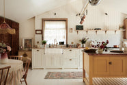 Looking straight towards the back wall of a bohemian-style kitchen that has tongue and groove panelled walls, solid floorboards, and a run of simple Shaker furniture, all painted in a creamy white shade. A mustardy yellow prep table sits in the centre of the room, creating a galley-style working area. The kitchen is fitted with soft pink marble worktops and styled with a collection of vintage pots and pans.