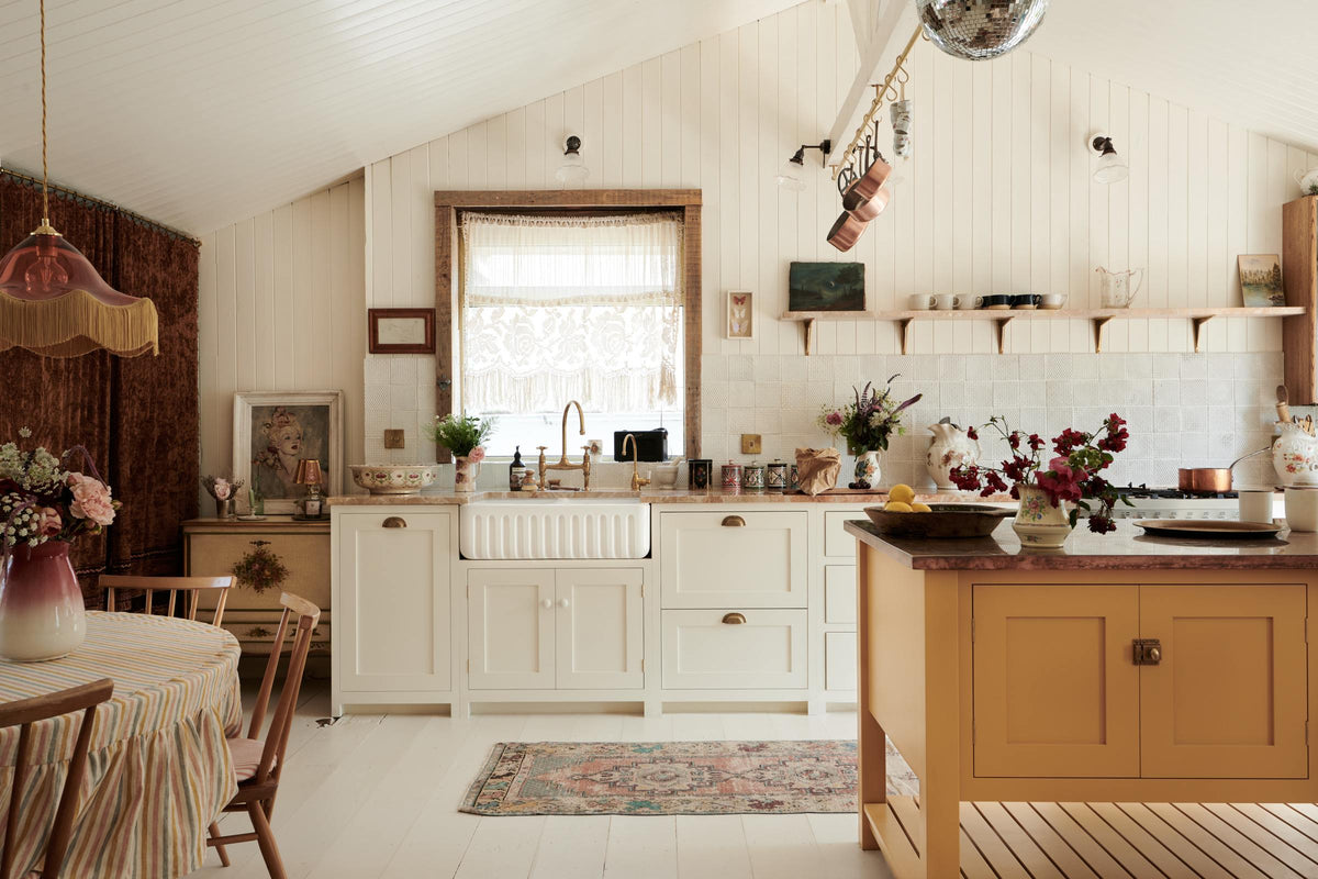 Looking straight towards the back wall of a bohemian-style kitchen that has tongue and groove panelled walls, solid floorboards, and a run of simple Shaker furniture, all painted in a creamy white shade. A mustardy yellow prep table sits in the centre of the room, creating a galley-style working area. The kitchen is fitted with soft pink marble worktops and styled with a collection of vintage pots and pans.