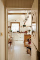 A view through the door reveals a bohemian-style kitchen that has tongue and groove panelled walls, solid floorboards, and a run of simple Shaker furniture, all painted in a creamy white shade. A mustardy yellow prep table sits at the centre of the room, creating a galley-style working area.