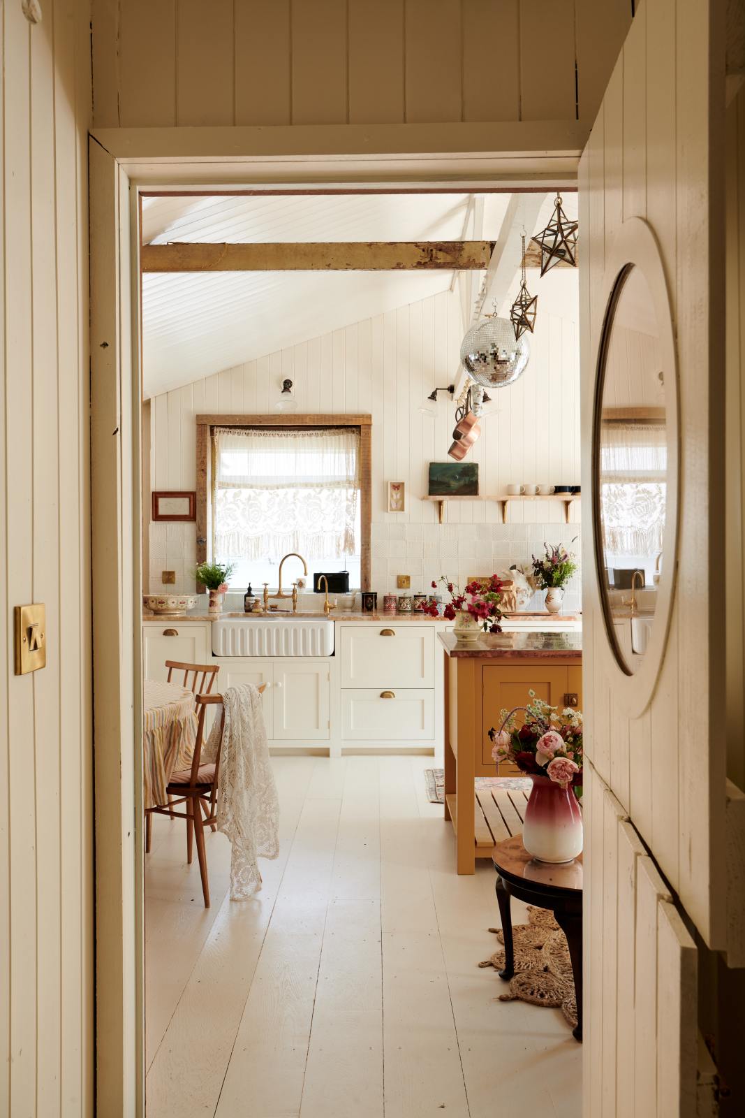 A view through the door reveals a bohemian-style kitchen that has tongue and groove panelled walls, solid floorboards, and a run of simple Shaker furniture, all painted in a creamy white shade. A mustardy yellow prep table sits at the centre of the room, creating a galley-style working area.