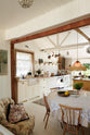 A beach house kitchen with Shaker cupboards, wooden floors and panelled walls, all painted in a fresh and bright off-white. There is a window behind the tactile ribbed sink and a brass mixer tap with a pretty patina. A table is just peeking into shot on the right-hand side, making this a very comfortable and social family kitchen.