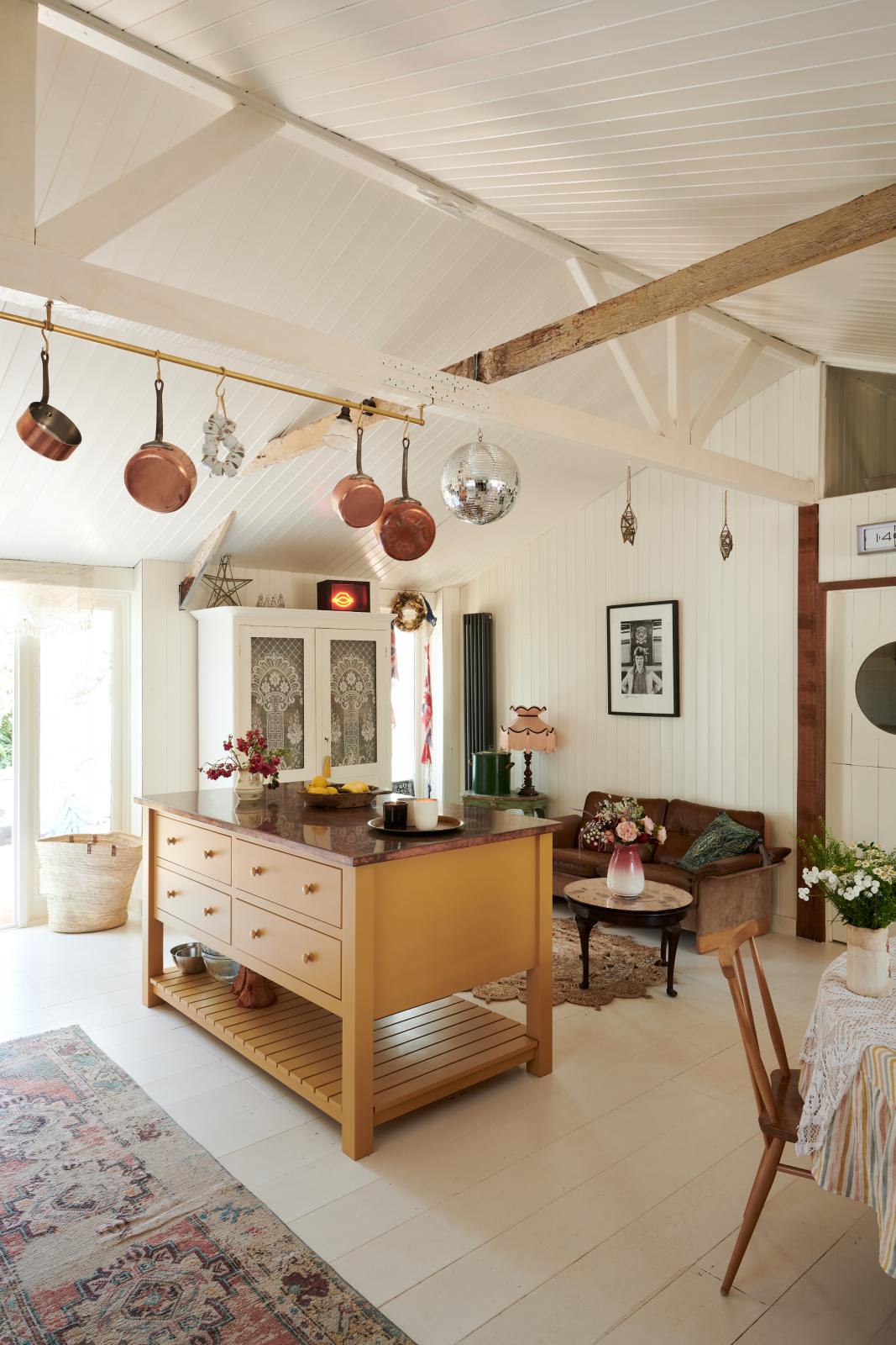 A photo of a bohemian-style kitchen. In the centre is a deVOL Shaker prep table, it has four drawers and a slatted base, it has been painted in a deep yellow and topped with aged copper. Behind is a sofa and a freestanding pantry. The walls and ceiling are panelled and painted in an off white, the floorboards are painted in a similar shade. There are painted beams running across the ceiling and a brass rail with copper pans.