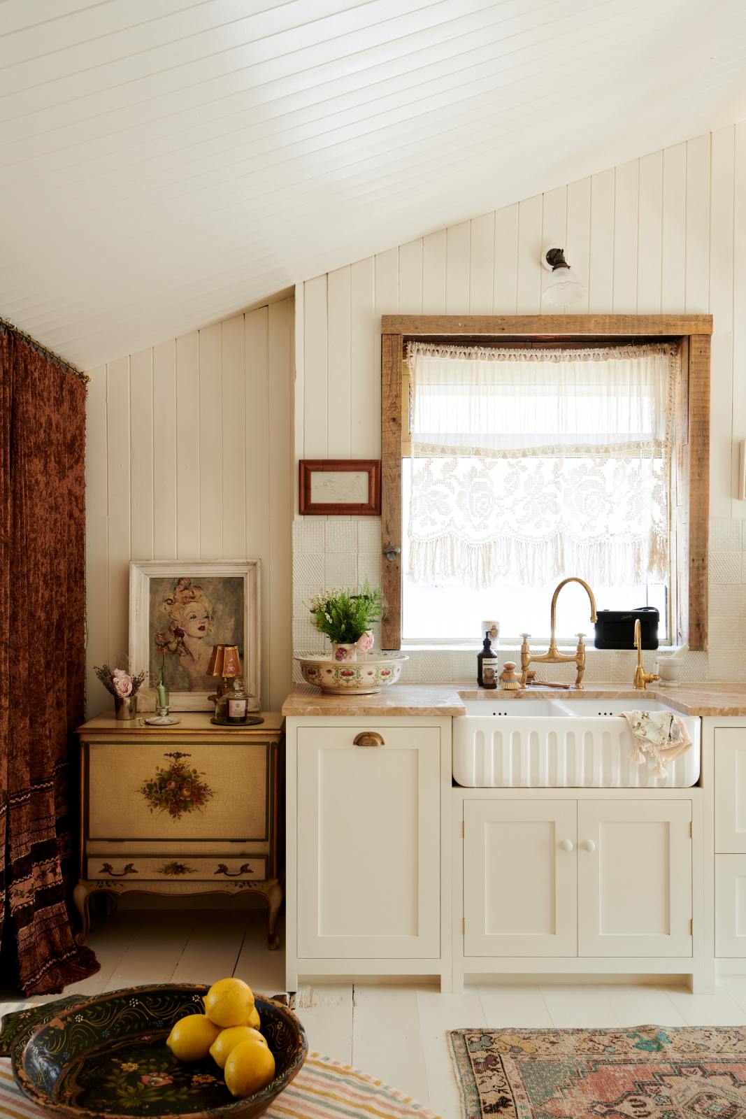 Looking straight towards the back wall of a bohemian-style kitchen that has tongue and groove panelled walls, solid floorboards, and a run of simple Shaker furniture, all painted in a creamy white shade. A mustardy yellow prep table sits in the centre of the room, creating a galley-style working area. The kitchen is fitted with soft pink marble worktops and styled with a collection of vintage pots and pans.