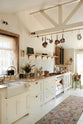 A diagonal view of a sink runs in a Bohemian-inspired beach house kitchen, featuring white cabinets with luminescent marble worktops and a fluted sink with two brass taps. An oriental-style carpet covers the floor and bronze pots in various sizes hang from the brass rail on the ceiling, enhancing a more vintage mood.