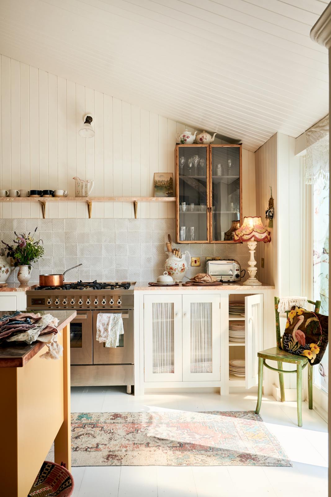 A straight-on view of a Bohemian-inspired beach house kitchen, featuring white cabinets with luminescent marble worktops. A beautiful antique-style lamp sits on the cabinet, and an oriental-style carpet covers the floor. A mustardy yellow prep table is placed at the centre of the room, while a green-painted wooden chair is positioned next to the window, adding a rustic touch.