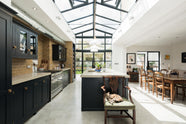 A bright and spacious pantry-blue Shaker kitchen under a wall-to-ceiling skylight features a long cooker run with wall cabinets, full-height cupboards, a kitchen island with a sink, a concrete worktop, and a tall bookshelf. Adorned with a grand wooden dining table with matching chairs, glass pendant lights, collected artworks, house plants, and a sleepy cat, it creates a warm and social space.