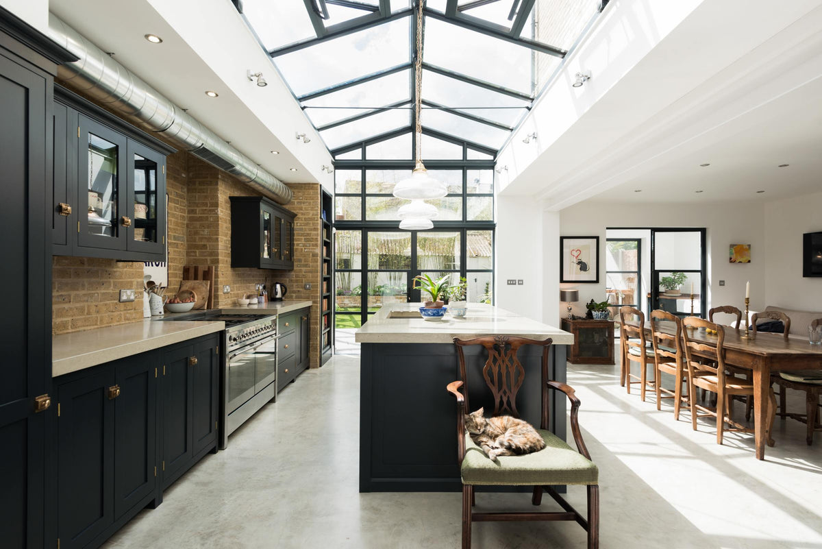 A bright and spacious pantry-blue Shaker kitchen under a wall-to-ceiling skylight features a long cooker run with wall cabinets, full-height cupboards, a kitchen island with a sink, a concrete worktop, and a tall bookshelf. Adorned with a grand wooden dining table with matching chairs, glass pendant lights, collected artworks, house plants, and a sleepy cat, it creates a warm and social space.