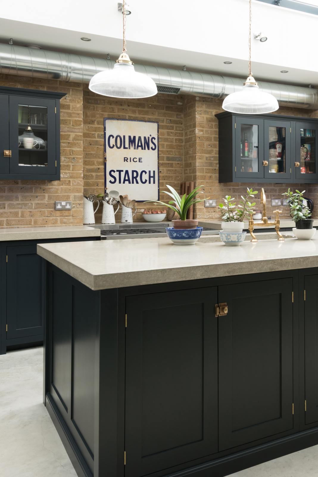 An angled view of a bright and spacious pantry-blue Shaker kitchen under a wall-to-ceiling skylight features a long cooker run with wall cabinets, a kitchen island with a sink, and a concrete worktop. Adorned with glass pendant lights, collected artworks, house plants, and everyday kitchen items, it creates a warm and social space.