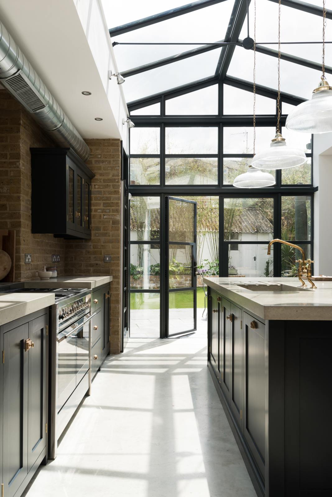 A bright and spacious pantry-blue Shaker kitchen under a wall-to-ceiling skylight features a long cooker run with wall cabinets, full-height cupboards, a kitchen island with a sink, a concrete worktop, and a tall bookshelf. Adorned with glass pendant lights, a muted pestle and mortar set, and chopping boards, it creates a warm and social space.