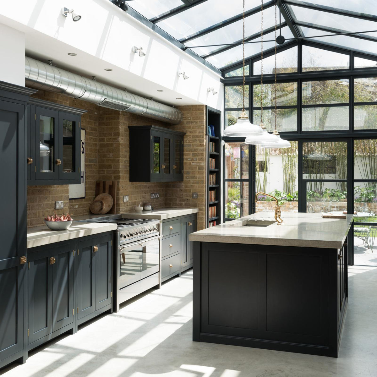 A bright and spacious pantry-blue Shaker kitchen under a wall-to-ceiling skylight features a long cooker run with wall cabinets, full-height cupboards, a kitchen island with a sink, a concrete worktop, and a tall bookshelf. Adorned with glass pendant lights, collected artworks, and a bowl of shallots, it creates a warm and social space.