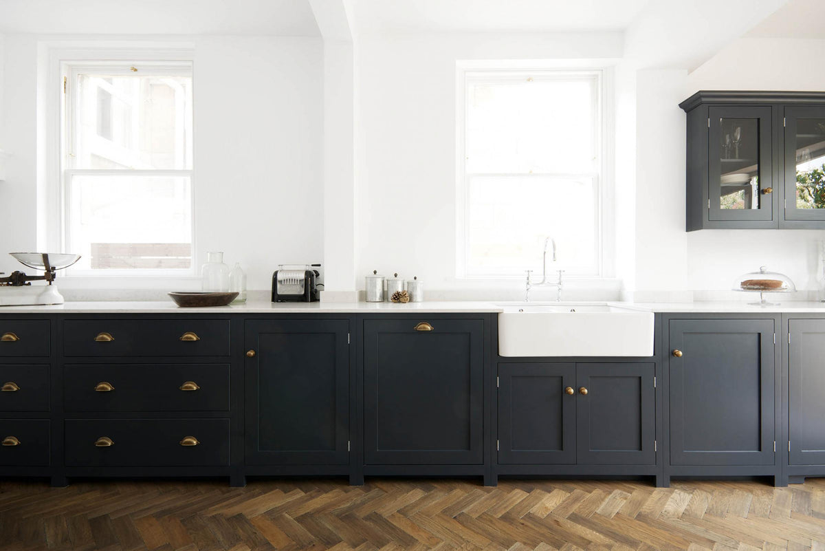 A bright and simple pantry-blue Shaker galley kitchen features a long sink and cooker run with a double Belfast sink, an antique silver Ionian tap, and glazed wall cupboards. Beautifully decorated with fundamental kitchen equipment such as a silver toaster, matching silver coffee, tea, and sugar containers, a glass cake stand and dome, a vintage scale, and reclaimed wooden parquet, it creates a stylish and effortless space.