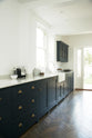 An oblique view of a bright and simple pantry-blue Shaker galley kitchen features a long sink and cooker run with a double Belfast sink, an antique silver Ionian tap, and glazed wall cupboards. Beautifully decorated with fundamental kitchen equipment such as a silver toaster, matching silver coffee, tea, and sugar containers, a glass cake stand and dome, and reclaimed wooden parquet, it creates a stylish and effortless space.