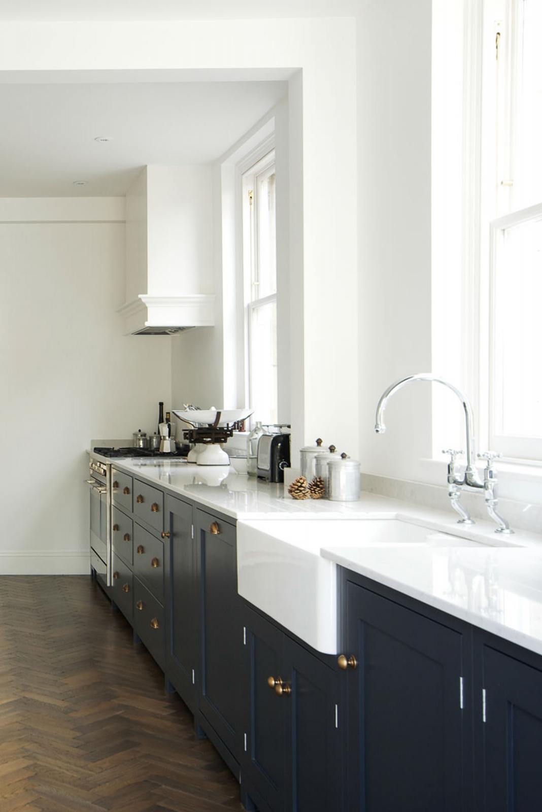 An oblique view of a bright and simple pantry-blue Shaker galley kitchen features a long sink and cooker run with a double Belfast sink, an antique silver Ionian tap, silver gas cooker, and a Shaker canopy. Beautifully decorated with fundamental kitchen equipment such as a silver toaster, matching silver coffee, tea, and sugar containers, a vintage scale, and reclaimed wooden parquet, it creates a stylish and effortless space.