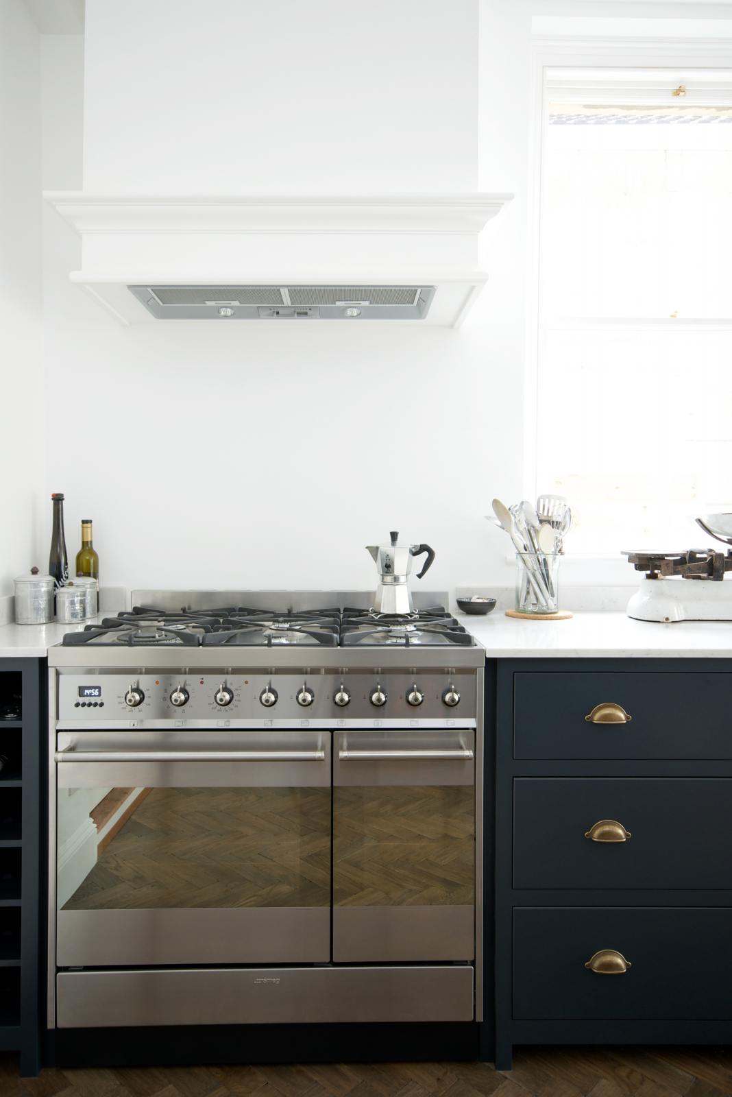 A bright and simple pantry-blue Shaker galley kitchen features a long sink and cooker run with a silver gas cooker and a Shaker canopy. Beautifully decorated with fundamental kitchen equipment such as a silver cafetiere, matching silver coffee, tea, and sugar containers, a vintage scale, and reclaimed wooden parquet, it creates a stylish and effortless space.