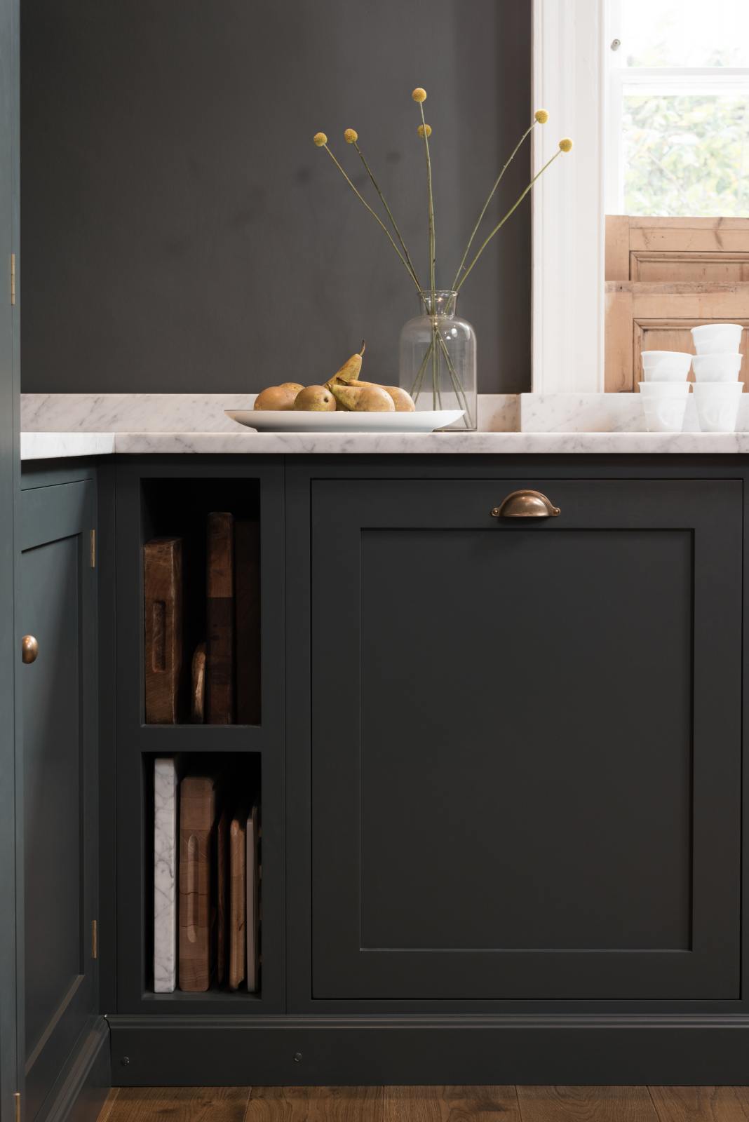 A quiet corner of a charcoal-black, classic shaker kitchen features a U-shaped sink and cooker run with Carrara marble worktop. Beautifully decorated with wooden chopping boards, charcuterie boards, plastic cups, a glass vase of flowers, fresh pears, and the original Georgian wooden sliders, the family kitchen strikes a wonderful balance between modern style and timeless character.
