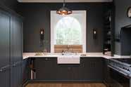 A charcoal-black, classic shaker kitchen features a U-shaped sink and cooker run with Carrara marble worktop, a chrome Ionian tap, open countertop cupboards, and a bank of fitted pantry cabinets. Beautifully decorated steampunk wall lights, a copper pendant light, fresh pears, and the original Georgian wooden sliders, the family kitchen strikes a wonderful balance between modern style and timeless character.