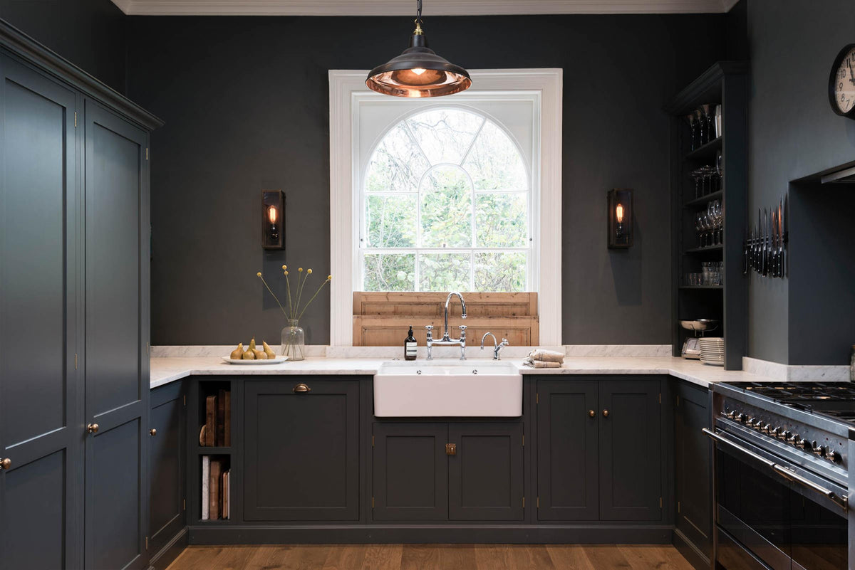 A charcoal-black, classic shaker kitchen features a U-shaped sink and cooker run with Carrara marble worktop, a chrome Ionian tap, open countertop cupboards, and a bank of fitted pantry cabinets. Beautifully decorated steampunk wall lights, a copper pendant light, fresh pears, and the original Georgian wooden sliders, the family kitchen strikes a wonderful balance between modern style and timeless character.