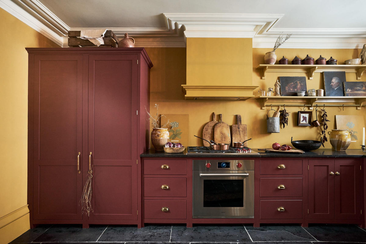 A busy and vibrant Refectory Red Shaker kitchen features an L-shaped sink and cooker run with a Belgian blue limestone worktop, a freestanding upright cupboard, and a classic Shaker canopy. Adorned with vintage items, beloved ceramics, still-life paintings, wooden chopping boards, red lettuce and deep red pomegranates, it creates a bright and wonderfully social kitchen space.