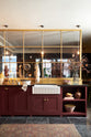A busy and vibrant Refectory Red Shaker kitchen features an L-shaped sink and cooker run with a Belgian blue limestone worktop, a single fluted Belfast sink, and aged brass Mayan taps. Adorned with vintage items, beloved ceramics, still-life paintings, dried chillies, red lettuce, and an arrangement of berry branches, creates a bright and wonderfully social kitchen space.