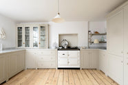 A minimalist, mushroom-coloured shaker kitchen features a cooker surrounding a chimney breast, a white gas stove, and a porcelain splashback paired with a marble worktop. It includes glazed cupboards, a shelving corner, and a spacious pantry cupboard.