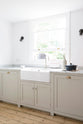 An angled view of a simple mushroom-coloured shaker kitchen features a marble worktop, a double Belfast sink with an antique silver Ionian tap, and glazed cabinets.