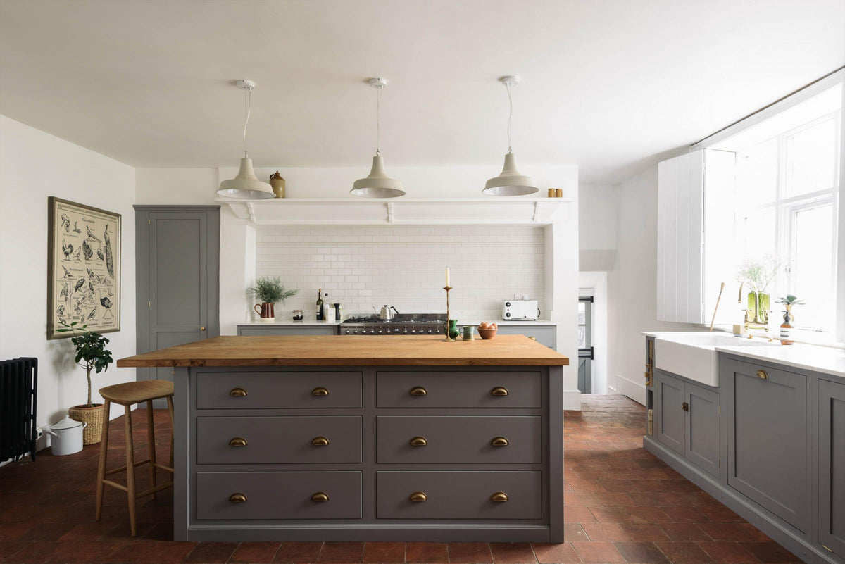 A simple, modern shaker kitchen in a historic Cheshire townhouse, featuring a cooker run, Silestone sink run, and a large island with a solid oak worktop and breakfast nook. Painted in muted cool grey with white walls and warm terracotta tile flooring. Finished with a Belfast sink, aged brass Ionian tap, and handcrafted ceramic bowls, jugs, and mugs.