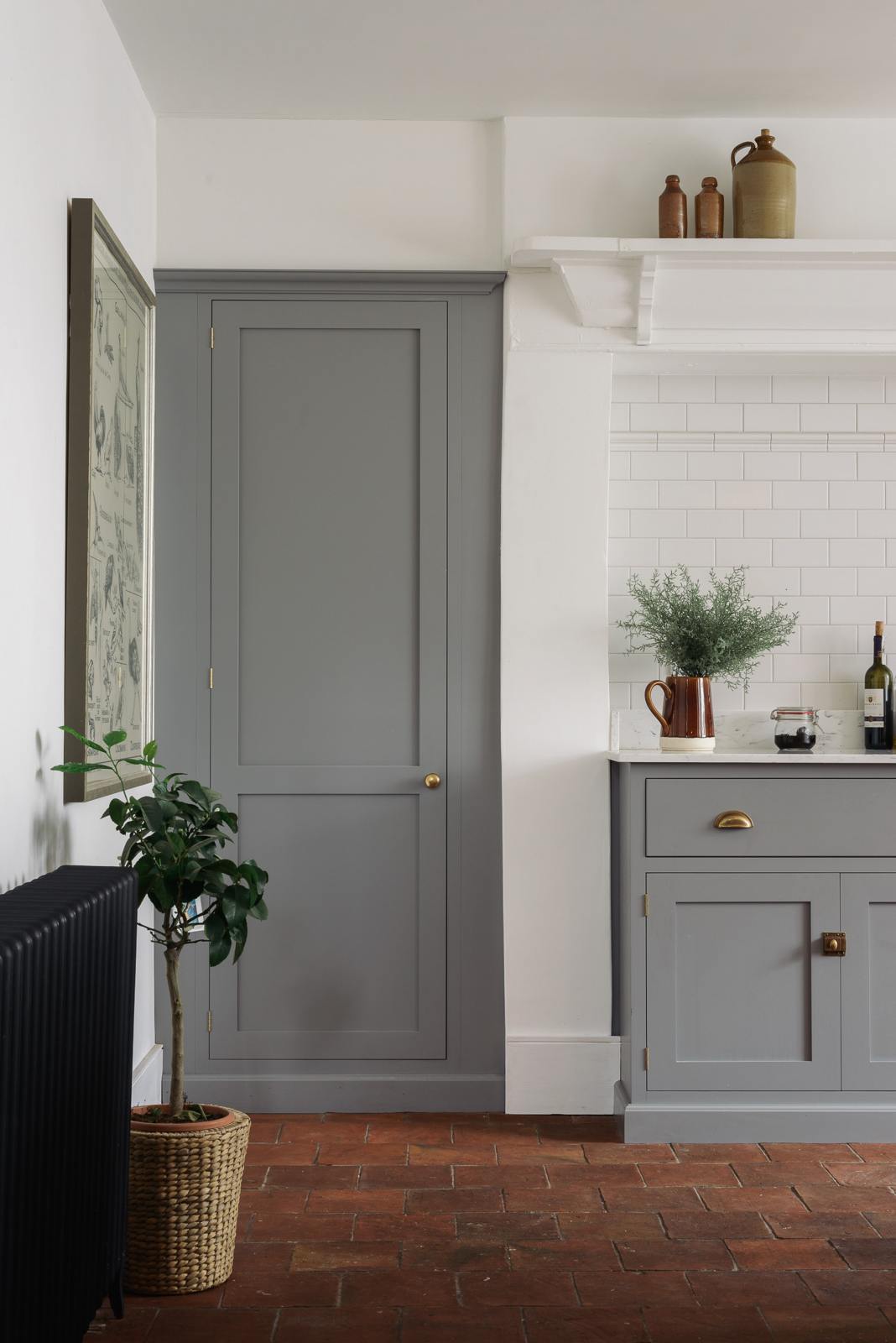 A closer look at the shaker kitchen’s full-height pantry door and the edge of a simple, modern cooker run, finished with white porcelain splashback tiles. Cupboards in mid-grey, terracotta tile flooring, and crisp white walls. Styled with a woven basket plant, a framed vintage illustration, and handcrafted ceramic bowls, jugs, and mugs.