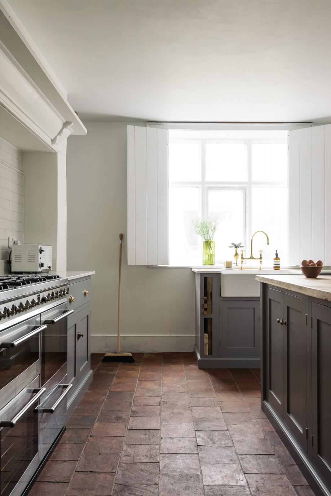 Looking between the simple, modern shaker kitchen’s cooker run and solid oak-topped island, facing the sink run with a Belfast sink and aged brass Ionian tap. Painted in muted cool grey with white walls, the warm terracotta tile floor glows in natural light from the open window.