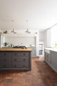 A closer look at a simple, modern shaker kitchen in a historic Cheshire townhouse, featuring a Silestone sink run with a Belfast sink and aged brass Ionian tap, and a large island with a solid oak worktop and a breakfast nook. Painted in muted cool grey with white walls and warm terracotta tile flooring.