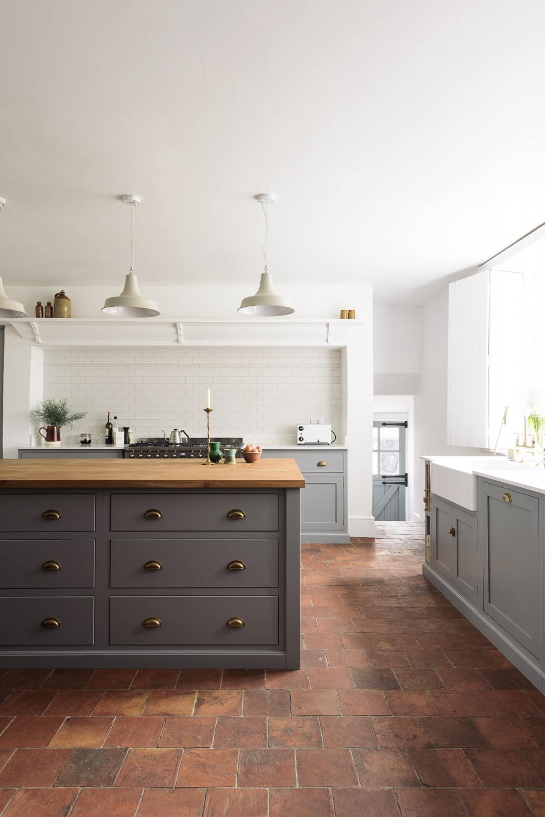A closer look at a simple, modern shaker kitchen in a historic Cheshire townhouse, featuring a Silestone sink run with a Belfast sink and aged brass Ionian tap, and a large island with a solid oak worktop and a breakfast nook. Painted in muted cool grey with white walls and warm terracotta tile flooring.