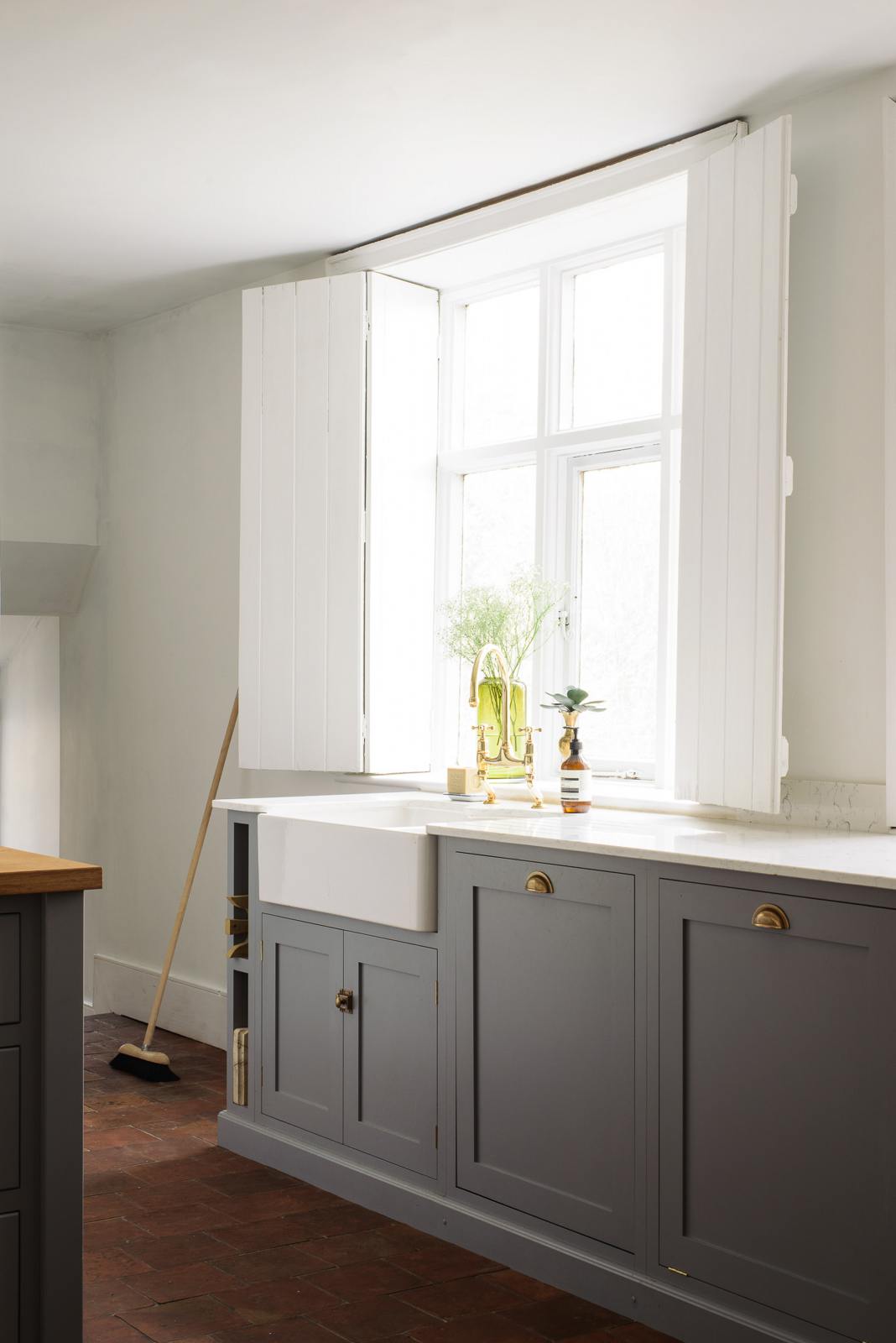 A close and angled view at a simple, modern shaker kitchen in a historic Cheshire townhouse, featuring a cooker run, Silestone sink run with a Belfast sink, aged brass Ionian tap, and a large island with a solid oak worktop and breakfast nook. Painted in muted cool grey with white walls and warm terracotta tile flooring.
