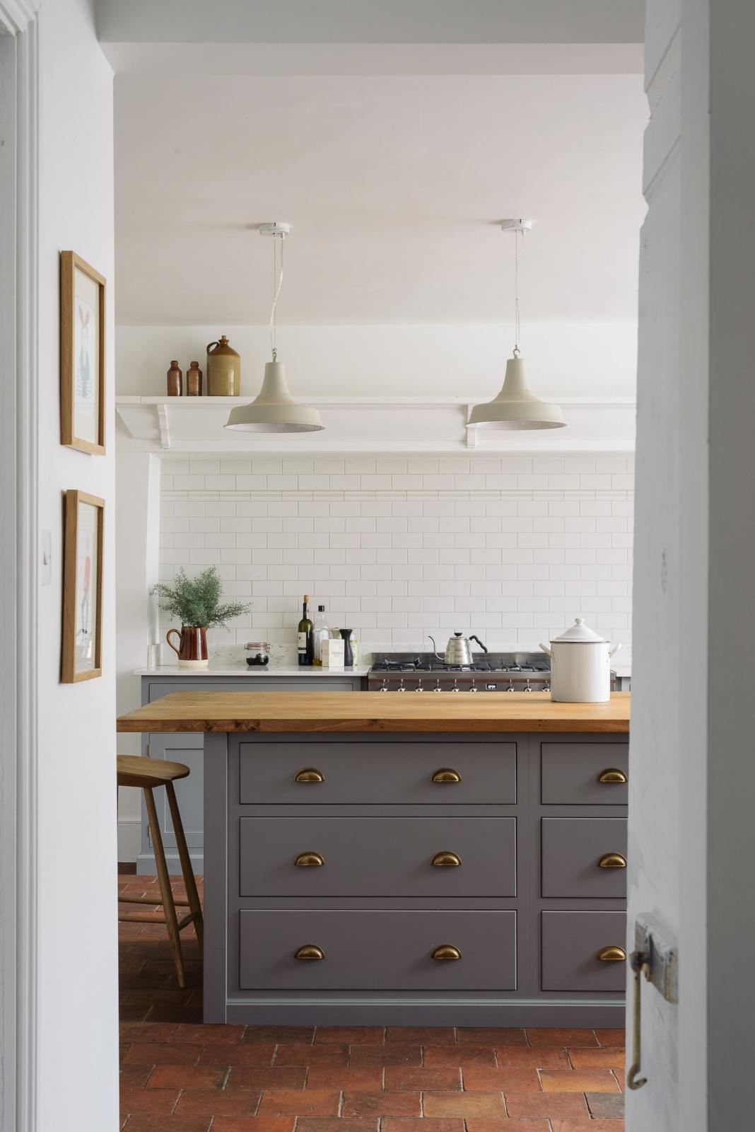 Looking through a narrow hallway, a simple, modern shaker kitchen in a historic Cheshire townhouse, featuring a Silestone worktop cooker run, and a large island with a solid oak worktop and a breakfast nook. Painted in muted cool grey with white walls and warm terracotta tile flooring.