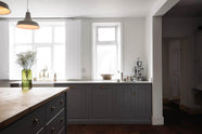 Bright light from the open windows highlights a simple, modern Shaker kitchen in a historic Cheshire townhouse. It features a cooker run, Silestone sink run with a Belfast sink and aged brass Ionian tap, plus a large island with a solid oak worktop and breakfast nook. Painted in muted cool grey with white walls and warm terracotta tile flooring.