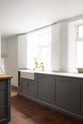 A close and angled view at a simple, modern shaker kitchen in a historic Cheshire townhouse, featuring a Silestone worktop sink run with a Belfast sink, aged brass Ionian tap, and a large island with a solid oak worktop and breakfast nook. Painted in muted cool grey with white walls and warm terracotta tile flooring.