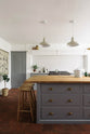 A close view at the solid oak bum stools tucked into the breakfast nook of a simple, modern Shaker kitchen island in a historic Cheshire townhouse. The kitchen features a cooker run with white porcelain splashback tiles, muted cool grey cupboards, white walls, and warm terracotta flooring.