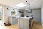 A spacious and contemporary stone grey Shaker kitchen features an L-shaped sink and cooker run with a Carrara worktop and splashback, a kitchen island, a canopy, and a bank of full-height cabinets. Elegantly decorated with glass-frilled pendant lights, vintage baskets, artisan ceramics, and deVOL bum stools, it creates a cool and inviting space.