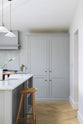A corner of a spacious and contemporary stone grey Shaker kitchen features an L-shaped sink and cooker run with a Carrara worktop and splashback, a canopy, a bank of full-height cabinets, and a kitchen island. Elegantly decorated with glass-frilled pendant lights, artisan ceramics, and a deVOL bum stool, it creates a cool and inviting space.