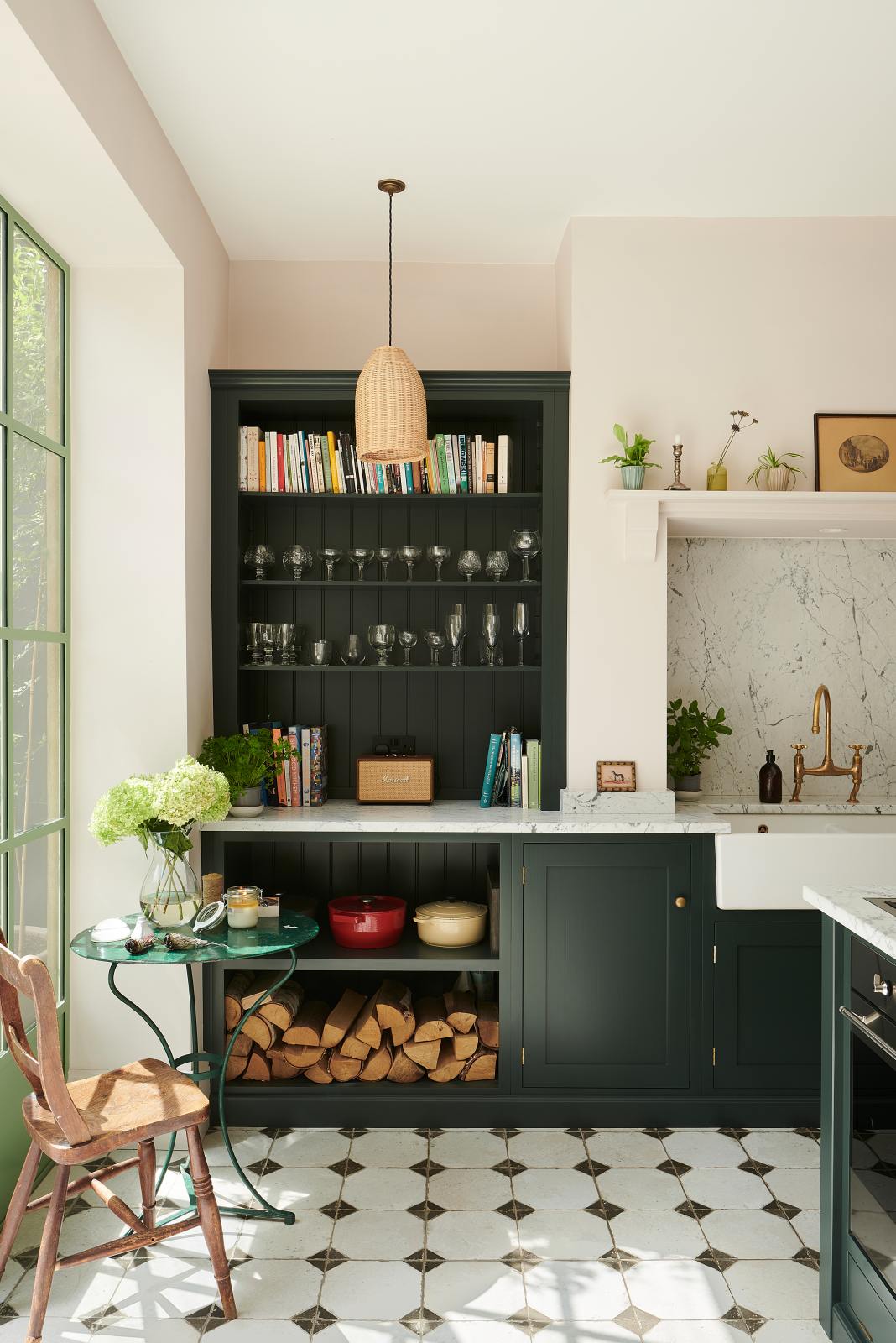 A detailed examination of the Bakehouse Green shaker kitchen’s sink run features a marble worktop, open cabinets and shelves, along with a double Belfast sink, an aged brass Ionian tap, and a Carrara marble splashback. Perfectly decorated with a small, deep green metal parlour table and a vintage wooden chair to add to a classic Mediterranean feel.