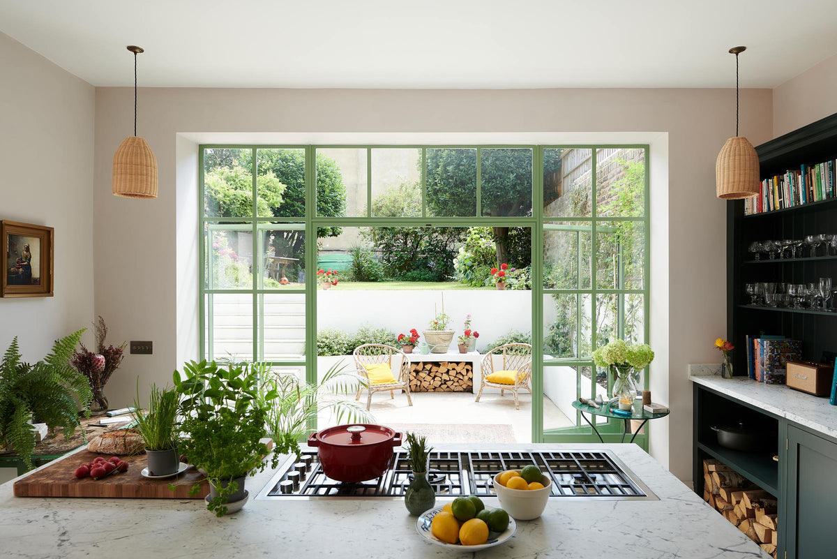 The Bakehouse Green shaker kitchen island features a beautiful Carrara marble worktop and a cooker. It overlooks wide Crittall windows painted in pistachio green, as well as a two-story patio and garden, creating a wonderful resort-like atmosphere right at home.